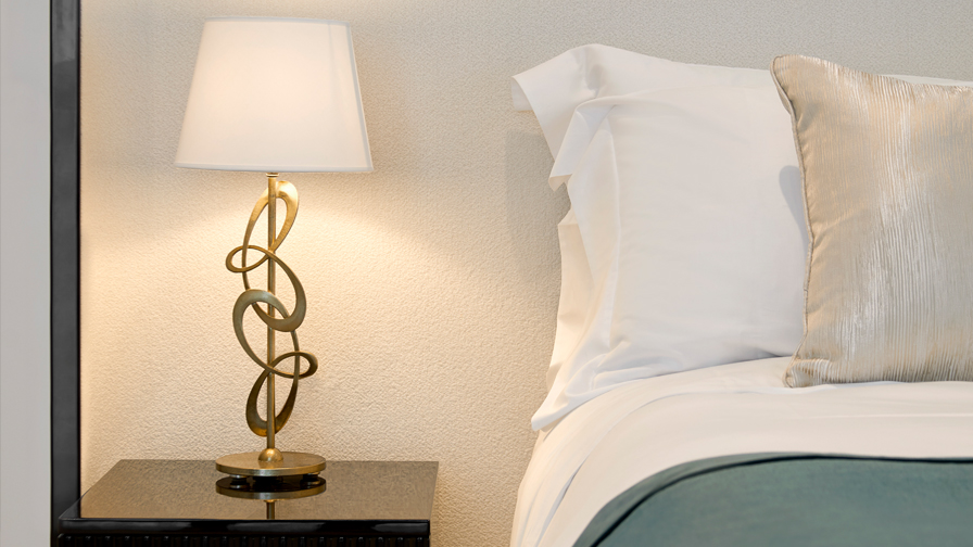 Close-up of luxury bedroom with white bedding, metallic accent pillow, and sculptural gold bedside lamp on a dark lacquered nightstand against a soft textured wall