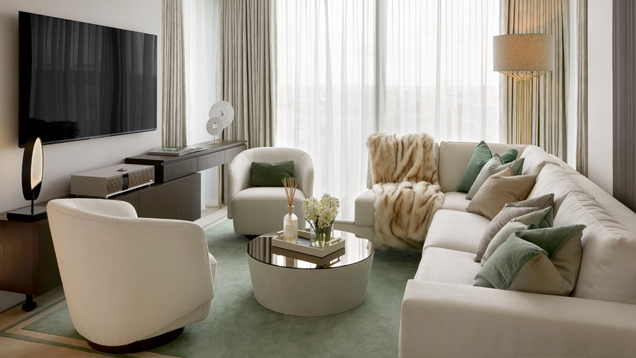 Contemporary luxury living room with ivory sectional sofa, velvet armchairs, soft green accents, fur throw, round mirrored coffee table, and full-height sheer curtains