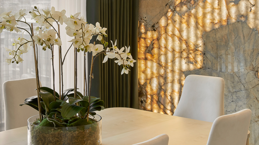 Elegant luxury dining area featuring white upholstered chairs, pale wood table, white orchid centerpiece, and a dramatic backlit onyx stone wall with green drapery