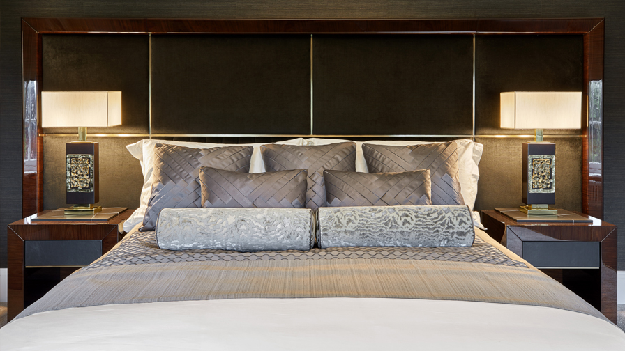 Close-up of luxury bed with oversized upholstered headboard, silk and velvet accent cushions, decorative bolster pillows, and designer bedside tables with statement lighting