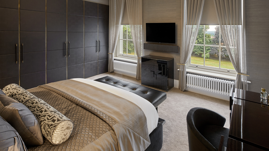 Luxury bedroom with bespoke upholstered bed, textured bedding, floor-to-ceiling wardrobes with gold detailing, and a sleek media unit below large sash windows with sheer curtains