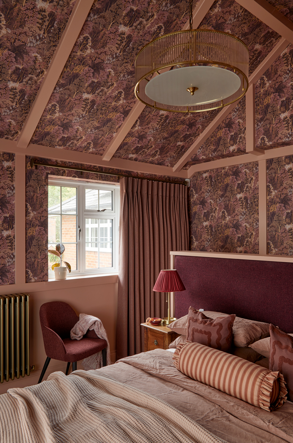 A cosy bedroom in deep pink and plum tones with patterned wallpaper across walls and ceiling. The bed has layered linens and cushions in warm shades, with a brass lamp, soft curtains, and a vintage-style pendant light adding warmth and texture.