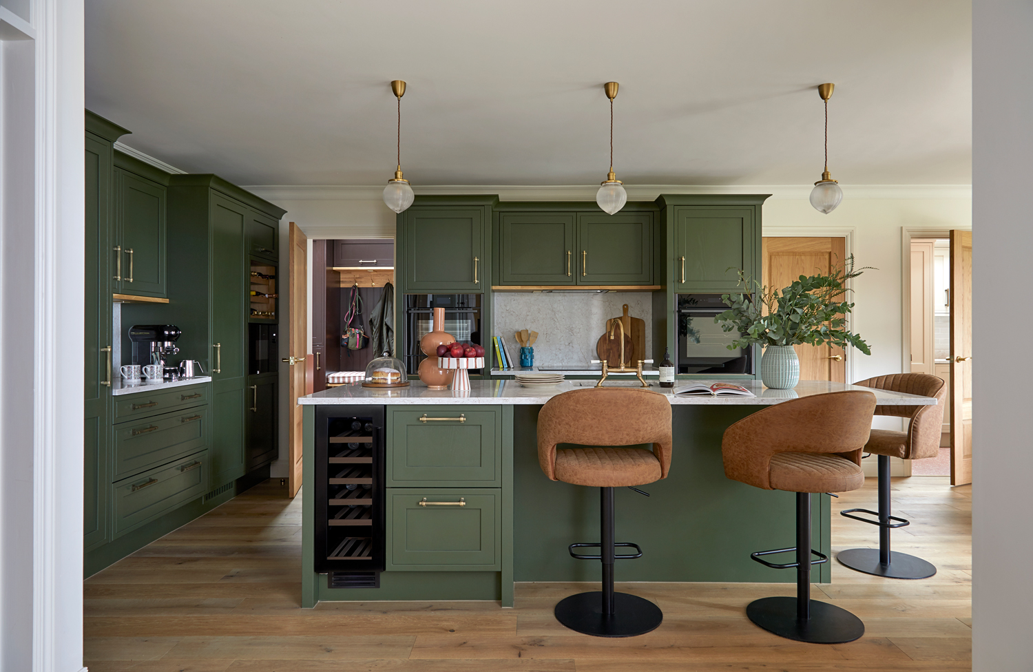 A welcoming kitchen with olive green cabinetry, brass hardware, and warm oak flooring. A marble-topped island with tan bar stools sits at the centre, complemented by globe pendant lights and soft brass accents that add warmth and balance.