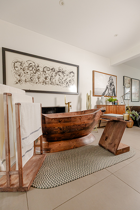 The Nautilus freestanding wooden bathtub displayed in an open-space cottage living room, with towel stand, artwork, and natural accents blending bath and home.