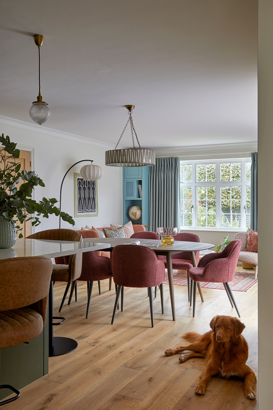 An open dining and living space with soft natural light, oak flooring, and warm tones. A wooden table is surrounded by deep red upholstered chairs, with a mix of vintage-style lighting and teal built-in shelving in the background. A golden retriever rests on the floor, adding a relaxed, homely feel.