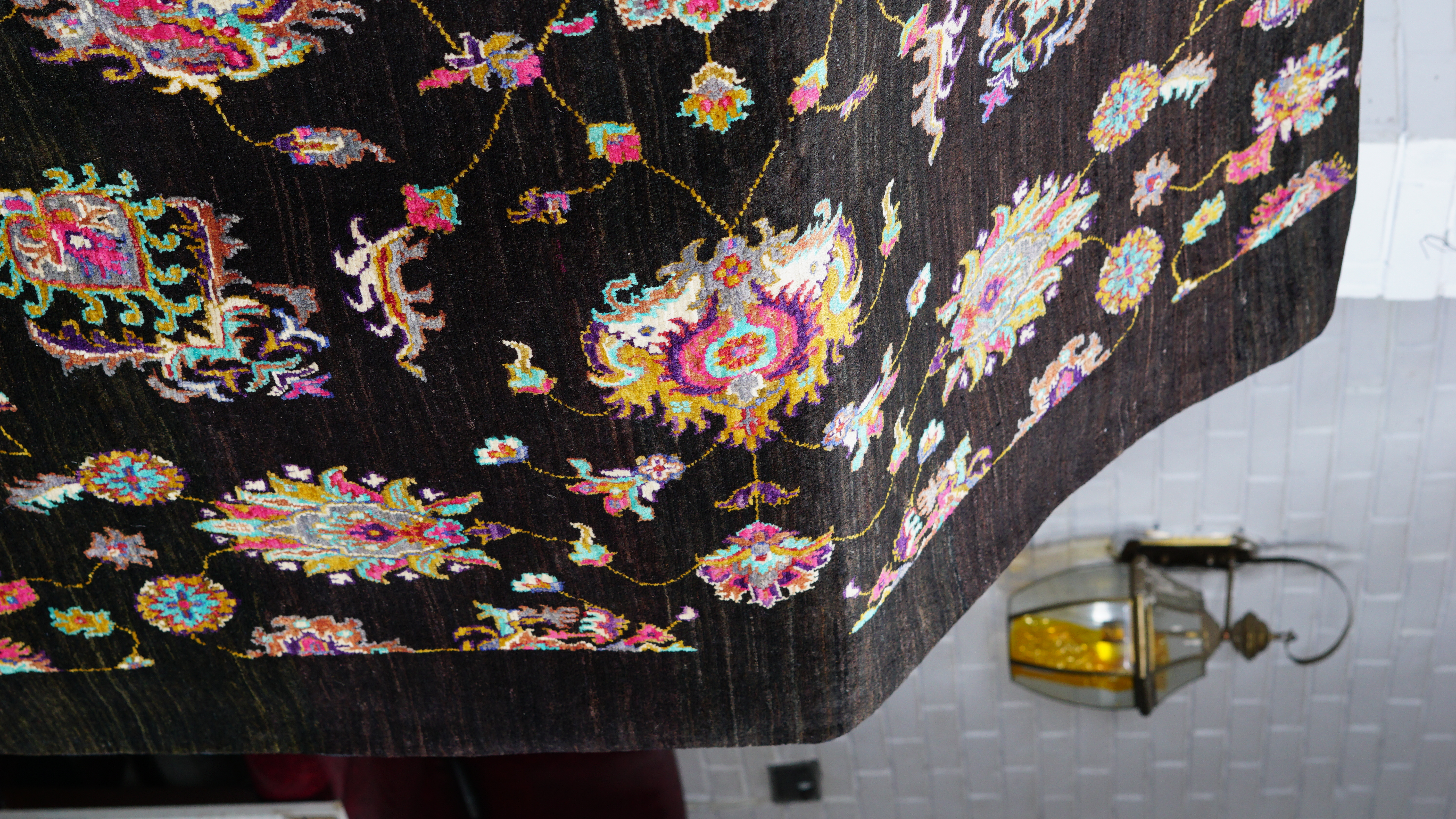 A close-up, angled view of a hand-knotted Afghan Kazak rug, featuring vibrant, colourful floral and geometric motifs on a dark brown field, draped over a surface.
