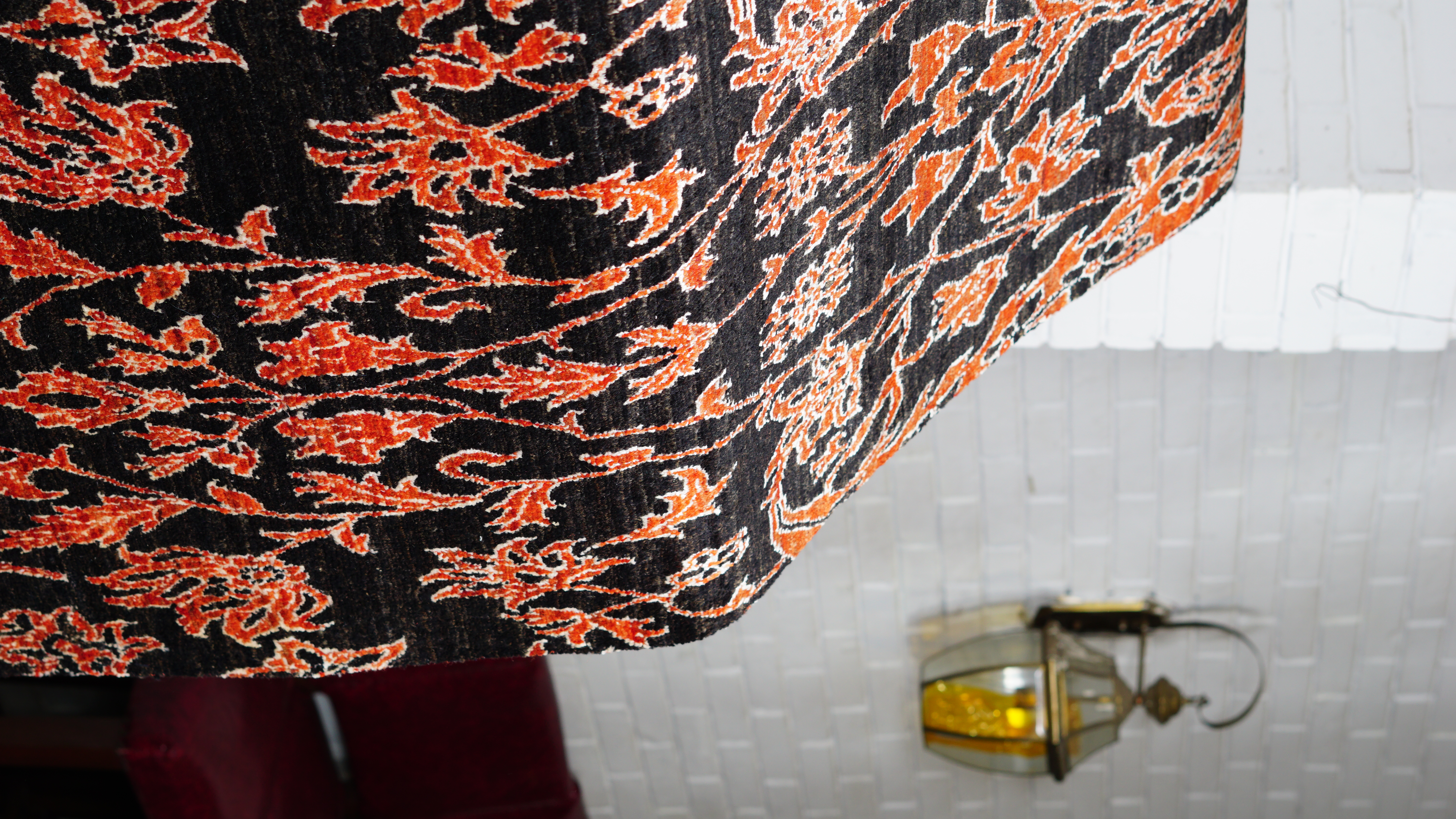 A close-up, angled view of a hand-knotted wool and silk rug, featuring an intricate, bright orange floral and vine pattern on a deep black background.