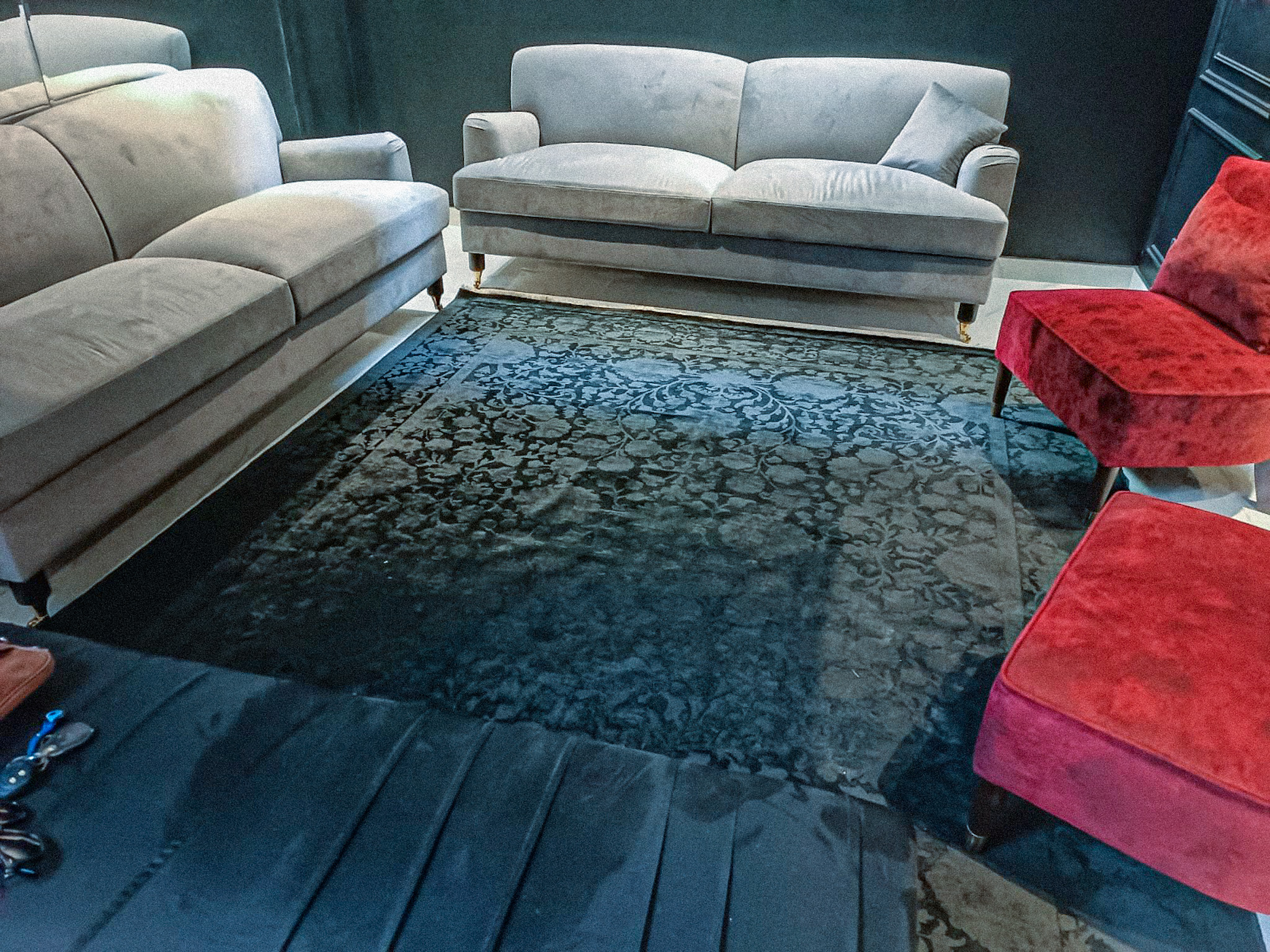 A dark, moody lounge featuring a black silk-wash rug with a subtle, tonal floral pattern, placed between grey velvet sofas and a red velvet chair.