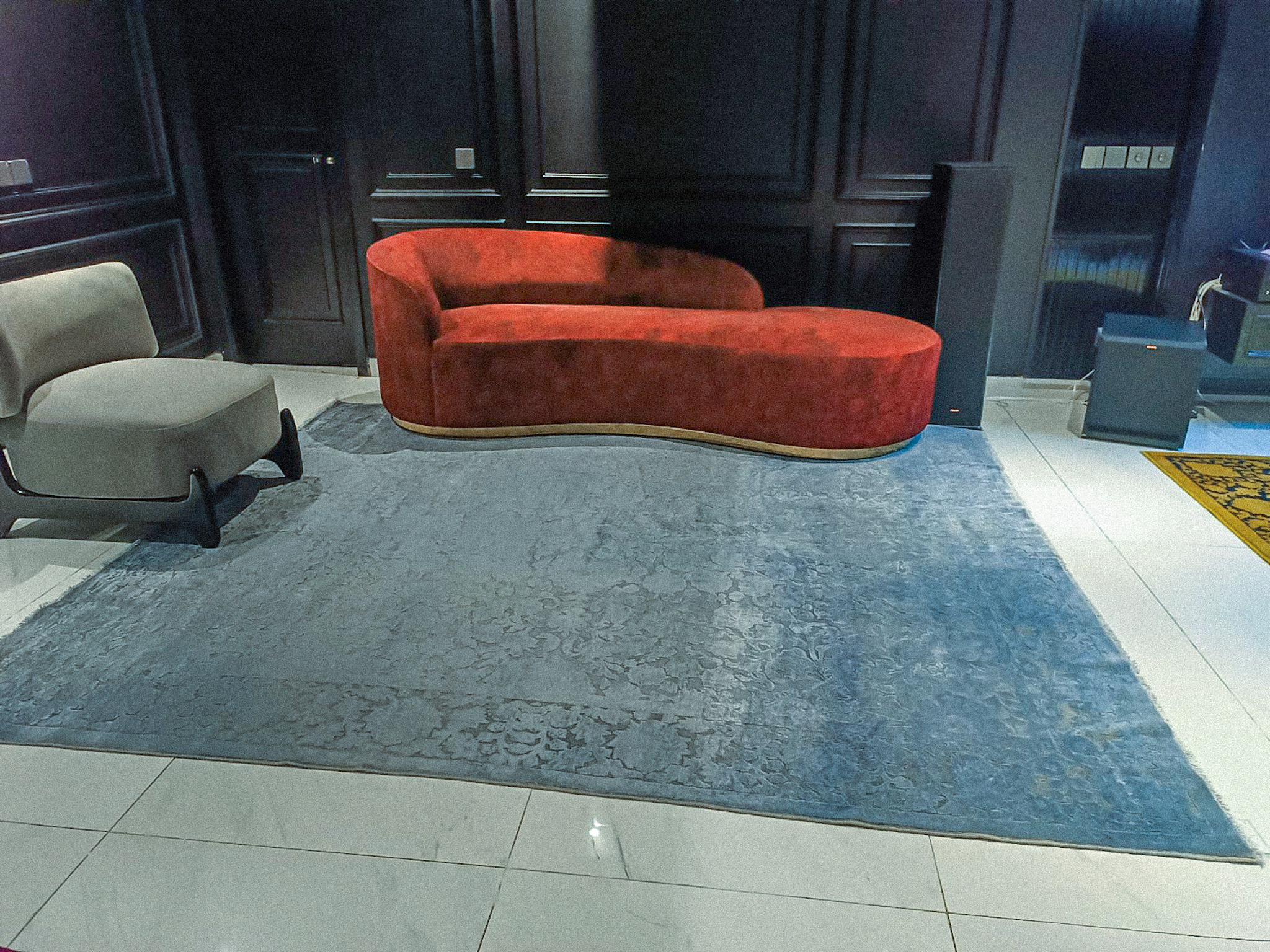 A large, silver-blue silk-wash rug with a tonal floral pattern anchors a modern lounge, featuring a striking, red-velvet curved chaise lounge and dark panelled walls.