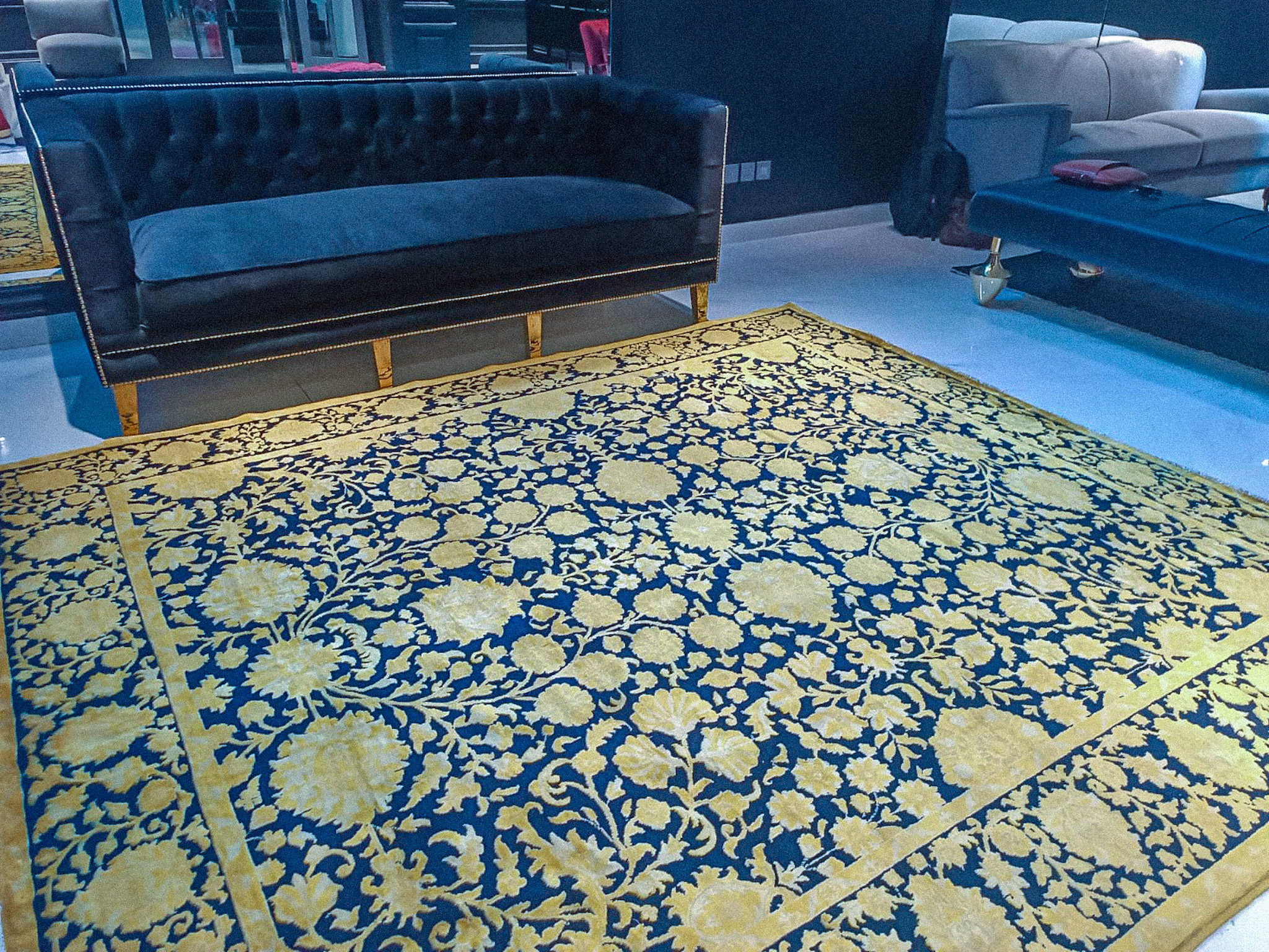 A hand-knotted, silk-wash rug with an opulent gold-yellow floral pattern on a deep blue background, placed in a modern showroom in front of a blue velvet sofa.