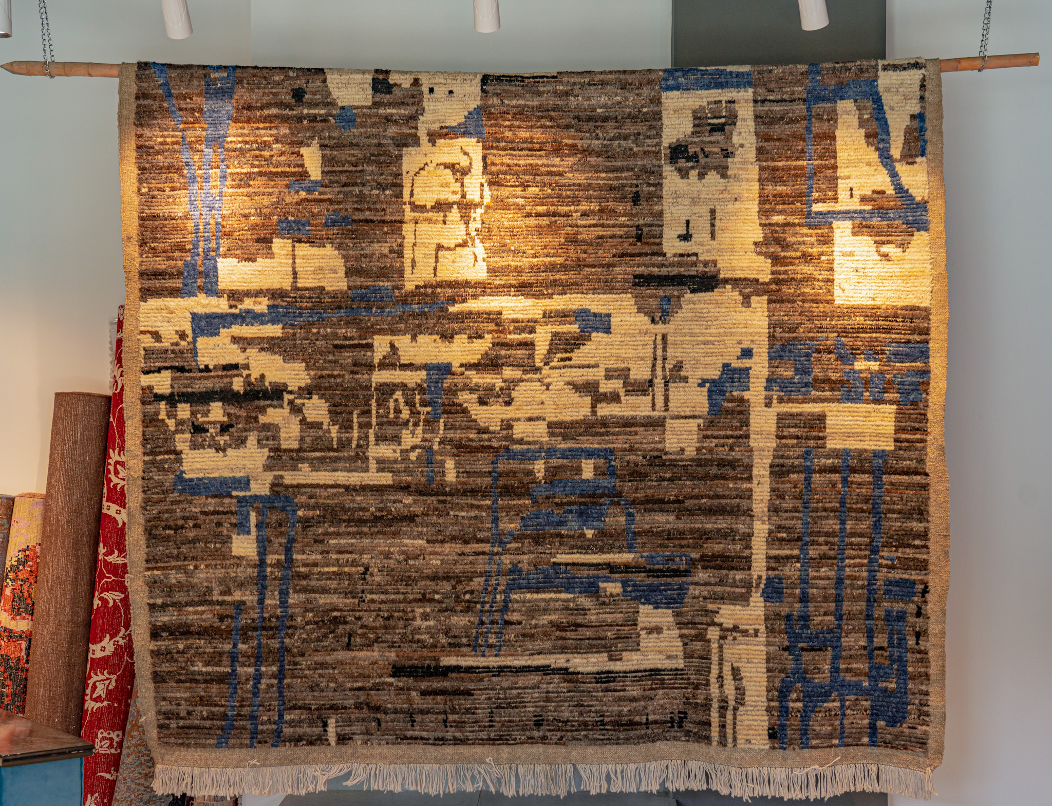 A large, hand-knotted, high-pile rug with a modern, abstract "deconstructed" pattern in brown, cream, and blue, hung for display against a white wall.
