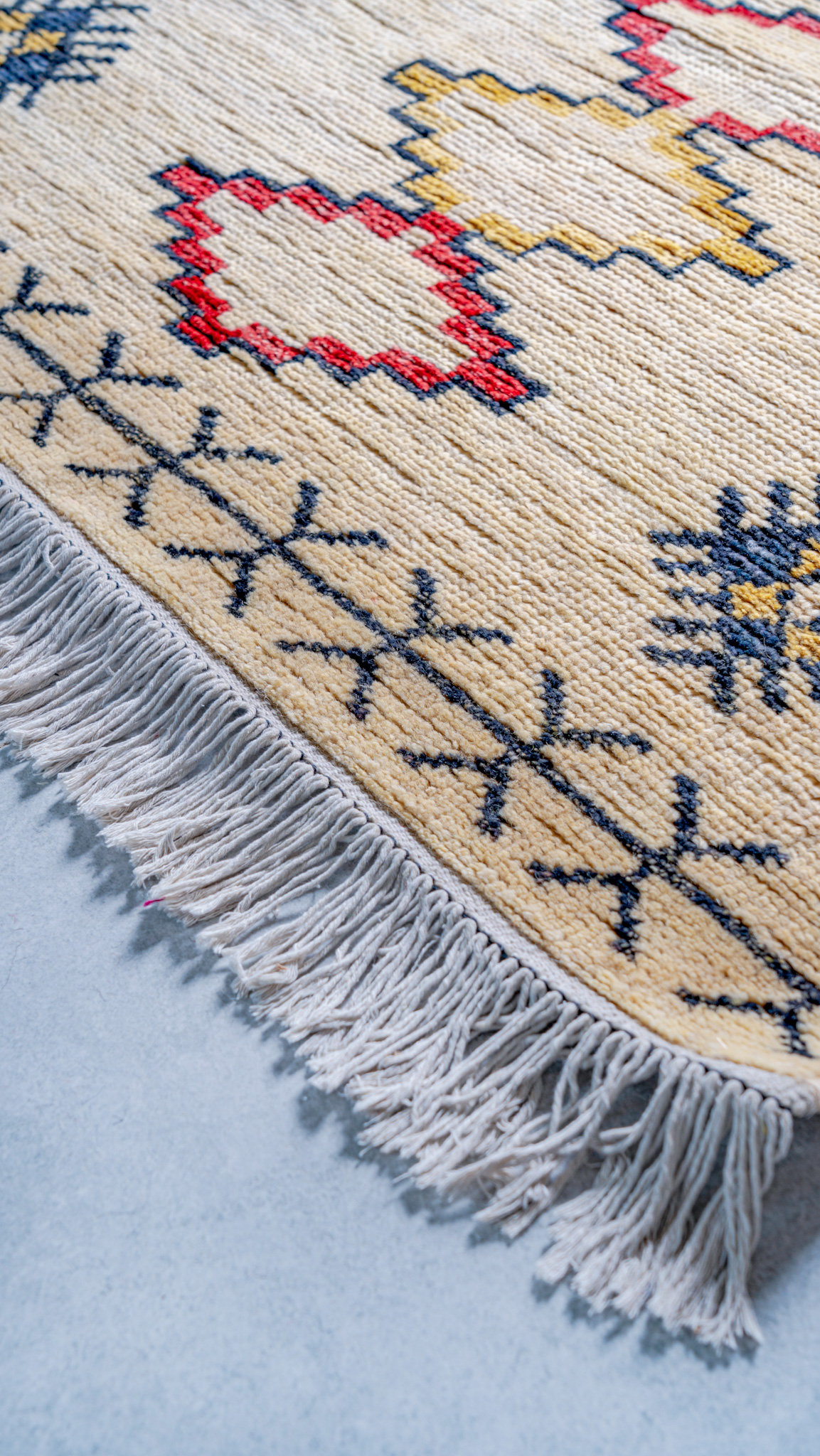 A close-up of the corner of a hand-knotted, cream-coloured rug, showing a geometric border in black and colourful motifs in red, yellow, and blue, with a white fringe.
