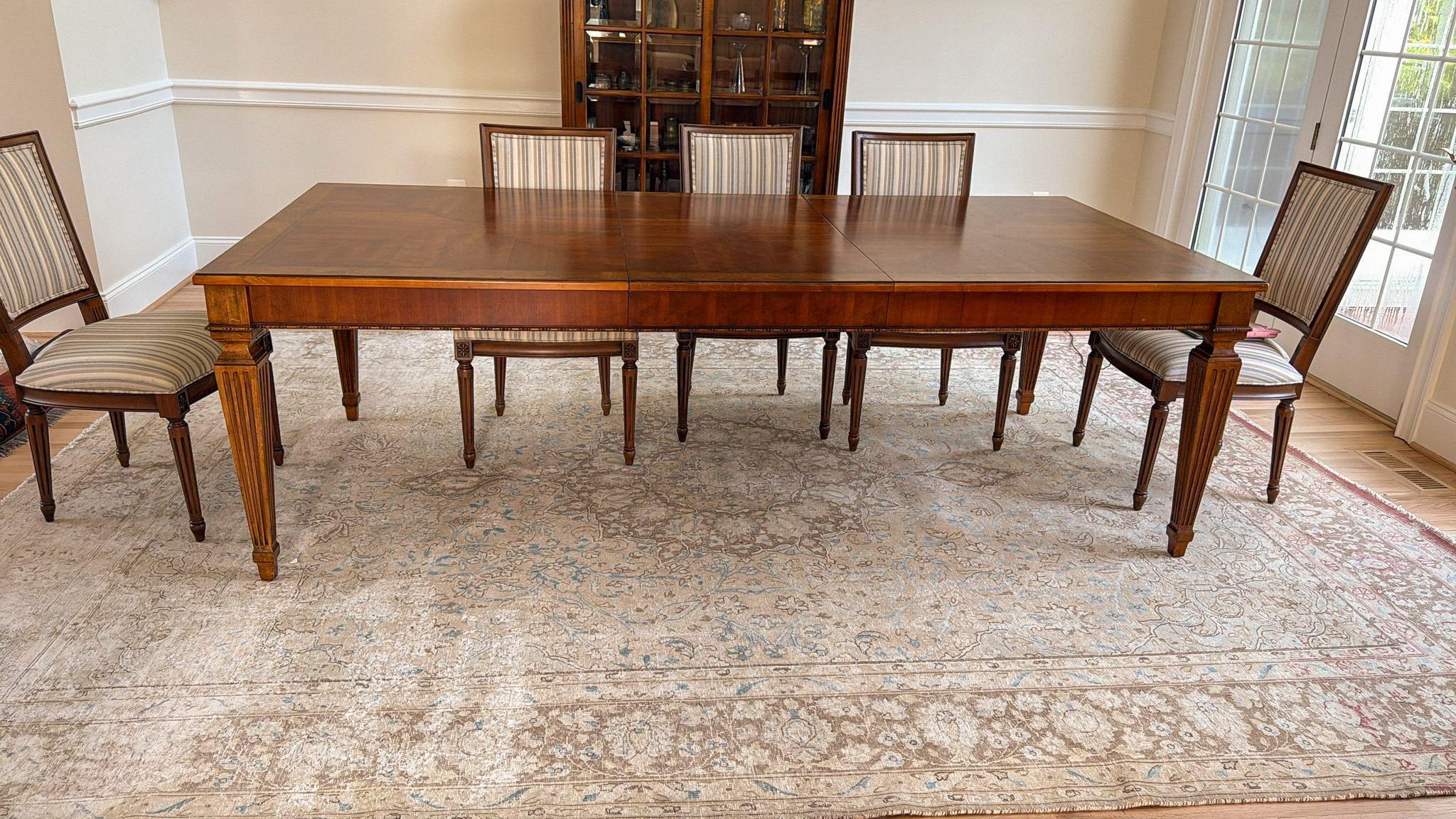 A large, overdyed Persian rug with a faded, distressed, vintage look in a light, neutral beige-grey, placed under a classic wooden dining table and chairs.