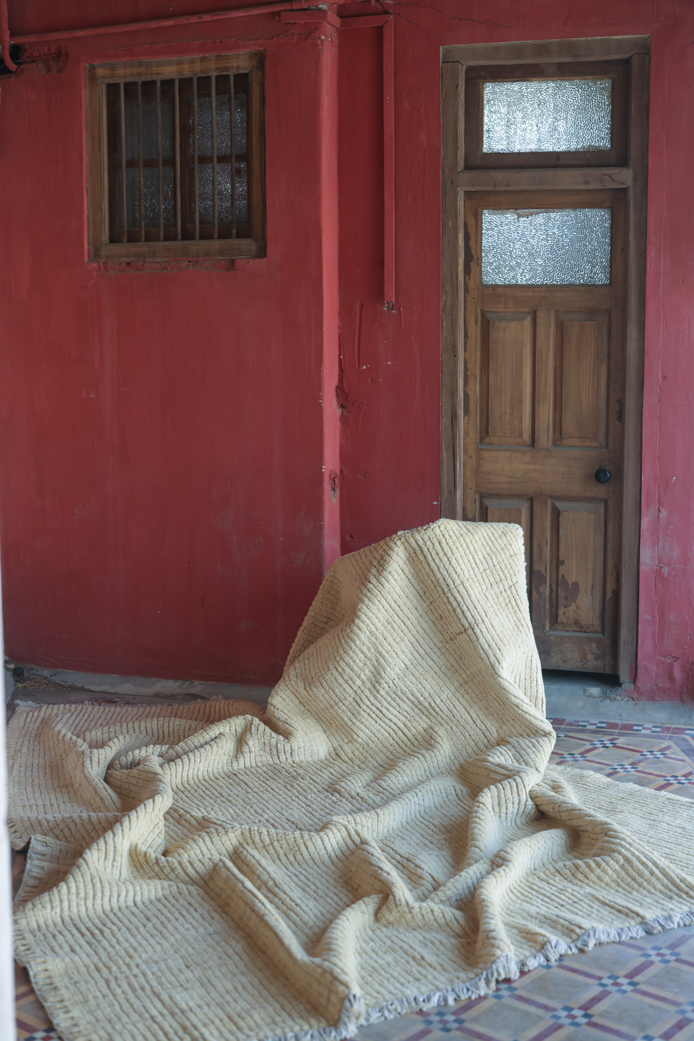 A close-up shot of a thick, hand-knotted rug from the Imaco Originals collection, featuring a textured, ribbed pattern in natural cream wool, draped over a surface on a patterned tile floor against a red wall.