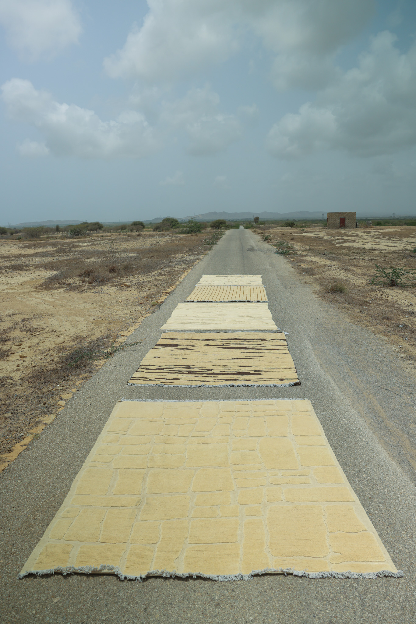 Several large, rectangular, hand-knotted rugs from the Imaco Originals Collection laid out on a long, straight road in a dry, open, natural landscape under a cloudy sky. The rug in the foreground has a high-low, sculpted geometric pattern.