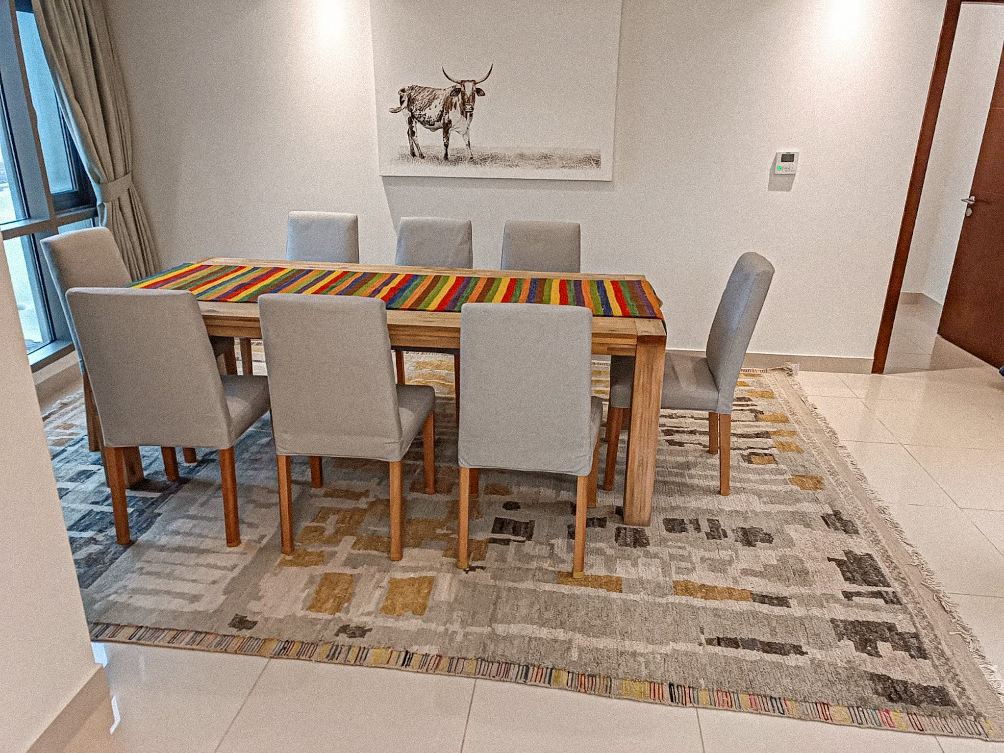 A modern dining room featuring a hand-knotted, low-pile rug with a geometric, abstract block pattern in grey, beige, and yellow, placed under a wooden dining table with grey chairs