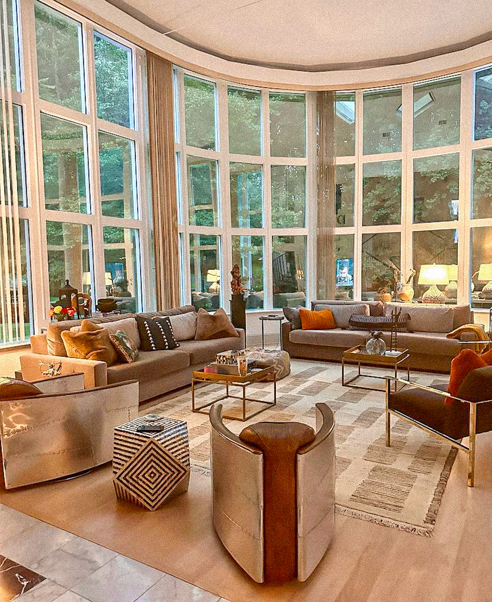 A grand, two-story living room with a curved wall of floor-to-ceiling windows, featuring a large, light-coloured, contemporary rug with a geometric block pattern that anchors the seating area.