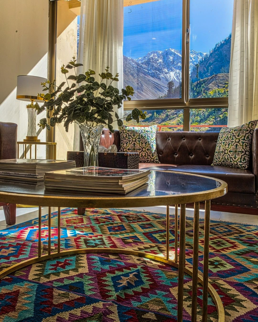 A close-up of a colourful, geometric kilim rug under a round, gold coffee table, with a brown leather sofa and a large window revealing a sunny, snow-capped mountain view in the background.