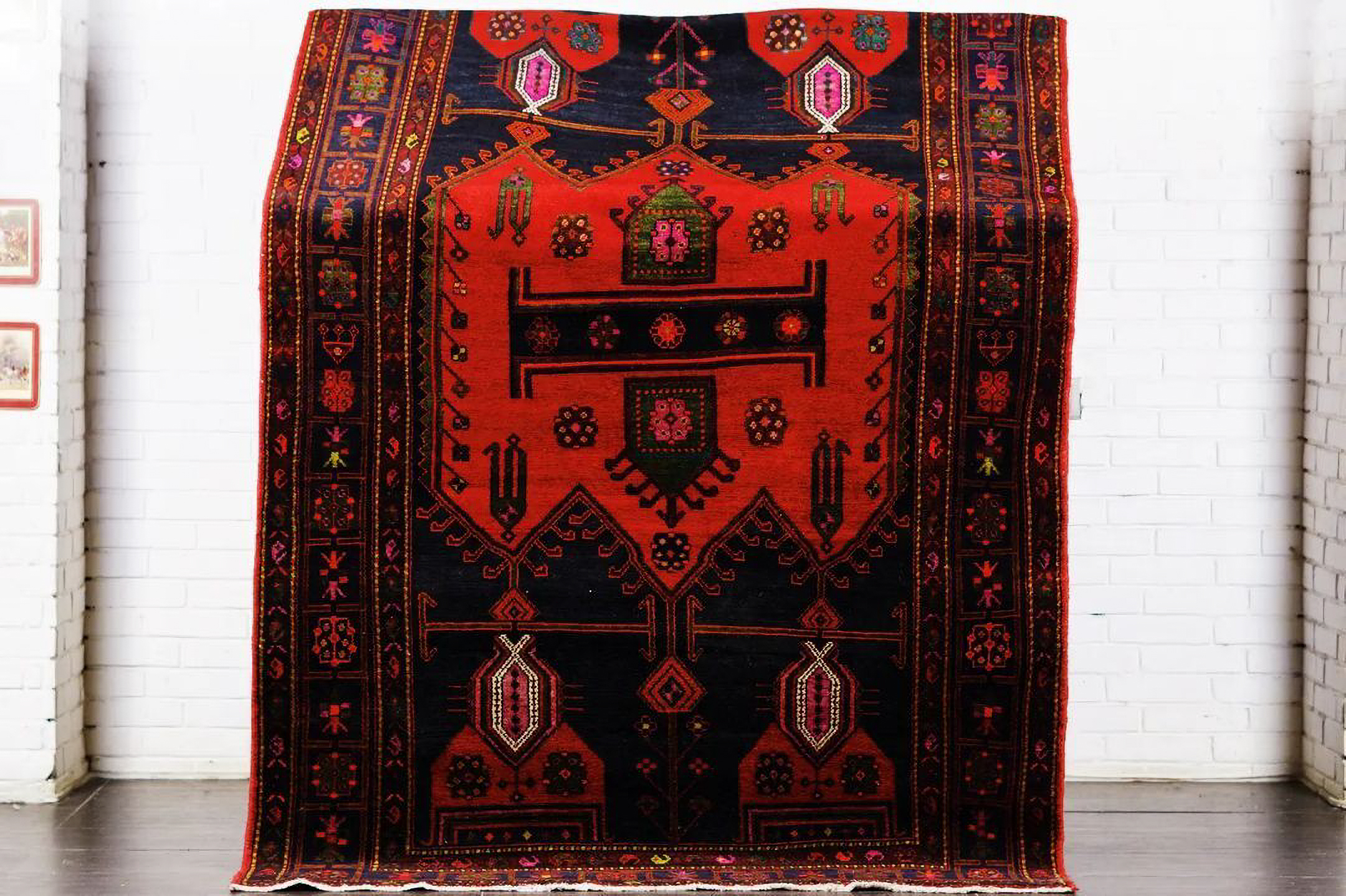 A hand-knotted Afghan tribal rug displayed vertically against a white brick wall, featuring a bold, geometric design with totemic motifs in red on a deep black field.