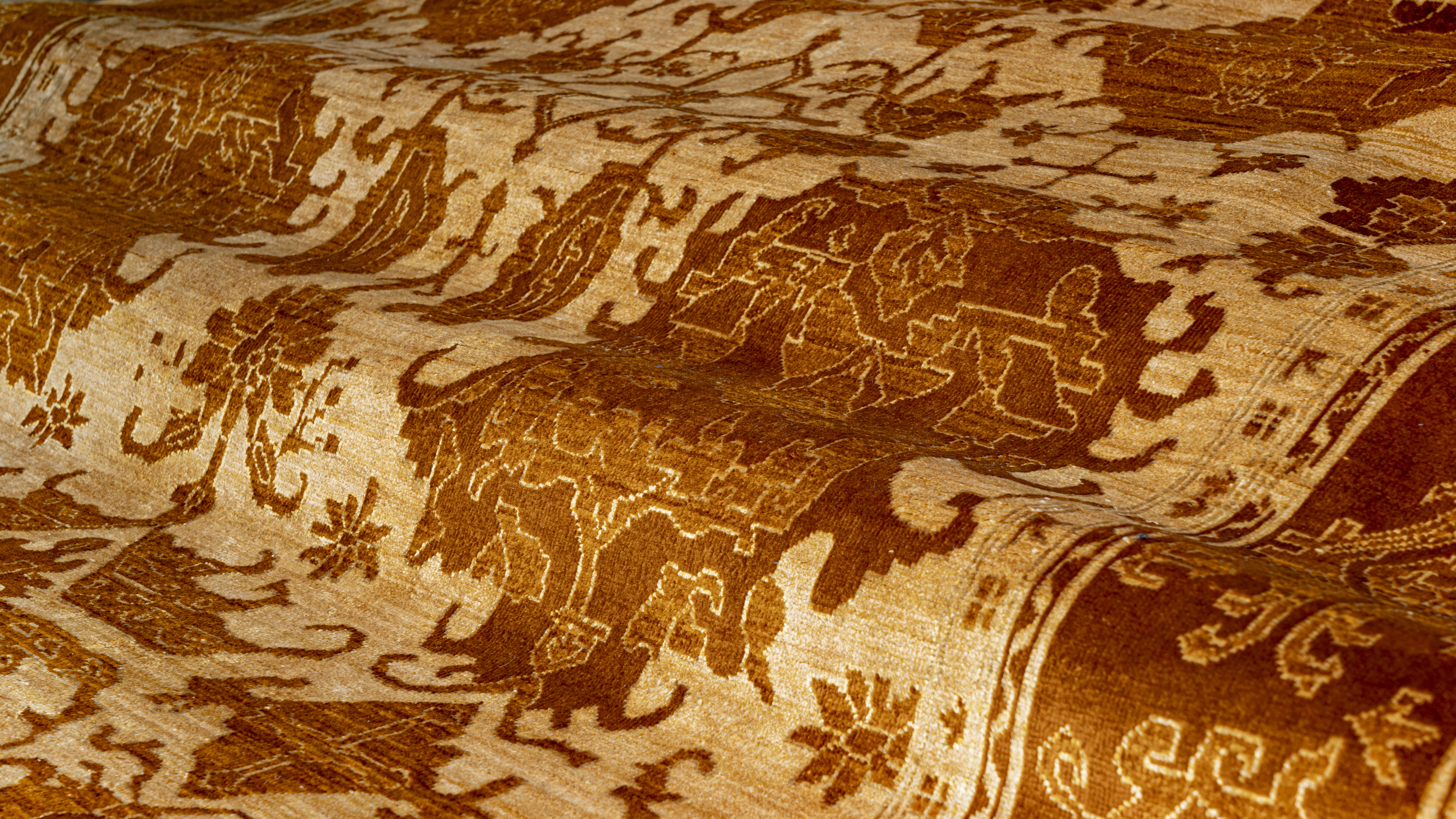 A close-up, angled view of a hand-knotted Afghan Ziegler rug, draped to show its texture, featuring an intricate, tonal pattern of rust-brown animal and floral motifs on a golden-beige field.