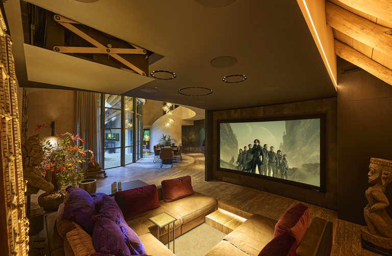 A wall-mounted large screen TV opposite a square sofa in a room with warm lighting and a spiral staircase in the back